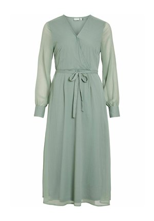 Wrapkjole i blødt, lys grøn stof. Har lange, gennemsigtige ærmer, et bånd i taljen og en svajende nederdel, der når til midt på lægen.