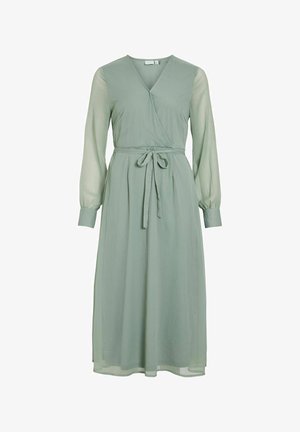 Wrapkjole i blødt, lys grøn stof. Har lange, gennemsigtige ærmer, et bånd i taljen og en svajende nederdel, der når til midt på lægen.