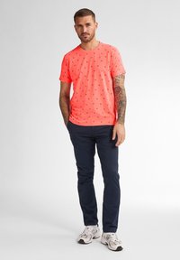 Neon-pinkes T-Shirt mit kleinen schwarzen Mustern, Rundhalsausschnitt. Kombiniert mit dunkelblauen Hosen und weißen Sneakers. Schlicher Hintergrund.
