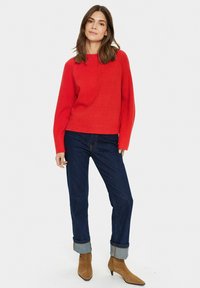 Maglione rosso a maglia con maniche lunghe, orlo a coste, abbinato a jeans blu scuro con risvolto e stivaletti beige a punta.