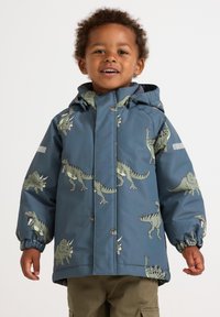 Un bambino in un giubbotto con cappuccio blu con motivi di dinosauri verdi, in piedi e sorridente su uno sfondo semplice.