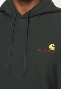 Felpa con cappuccio verde scuro dal morbido texture, con un logo ricamato giallo e bordeaux. Include cordini di regolazione e tasca a marsupio.