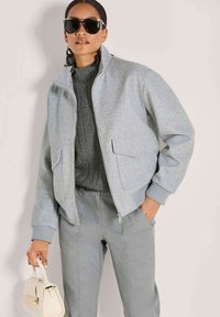 Frau trägt eine graue Zip-Up-Jacke, einen grauen Pullover, passend graue Hose, dunkle Sonnenbrille, große Ohrringe und trägt eine kleine weiße Handtasche.