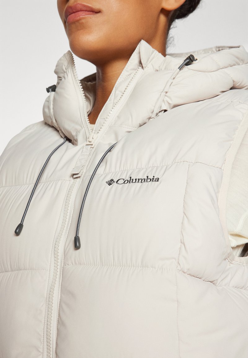 Close-up van een persoon die een lichtbeige Columbia puffer vest draagt met een hoge kraag en verstelbare koordjes.