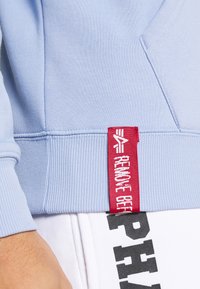 Polsino di felpa azzurro chiaro con un'etichetta di stoffa rossa che riporta "RIMUOVERE PRIMA" attaccata sopra pantaloni bianchi con scritte nere parziali.