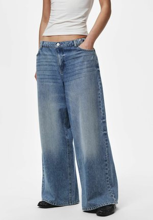 Personne portant un jean bleu délavé à jambes larges, les mains dans les poches et un débardeur court blanc, debout sur fond blanc.