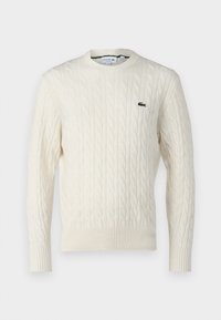 Cream cable-knit sweater with a round neckline, ribbed cuffs and hem, and a green Lacoste logo on the chest. Textured fabric enhances detail.