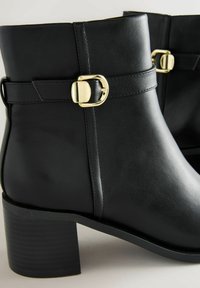 Mustad nahkmatid pahkluu saapad ümarate varvastega, külgmise pandla detailiga ja tekstureeritud plokk-kandiga. Sile viimistlus minimaalsete õmblusaktsentidega.