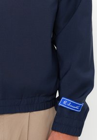 Marineblauwe windjack met elastische zoom en manchetten. Bevat een prominent blauw label met witte tekst op de mouw. Gladde stofstructuur.