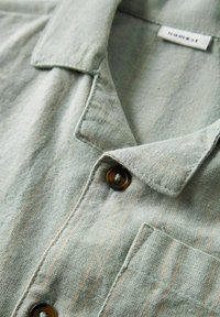 Camicia di lino verde chiaro con strisce arancioni verticali, dotata di colletto a ribaltina e un singolo bottone. Include un dettaglio di taschino sul petto.