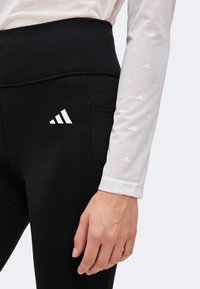 Femme portant des leggings noirs taille haute avec un logo blanc Adidas près de la hanche et un haut blanc à manches longues avec de petits logos Adidas.