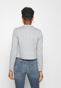 Harmaa lyhythihainen takki pitkillä hihoilla, jossa on pehmeä pinta ja yksinkertainen kaulus, yhdistettynä sinisiin kapeisiin farkkuihin.