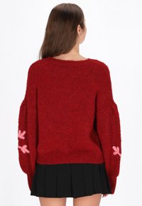 Roter Strickpullover mit lockerem Schnitt, langen Ärmeln und rosa Schleifendetails an den Bündchen. Kombiniert mit einem schwarzen Faltenrock.