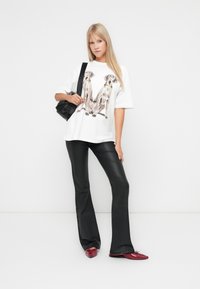 Witte oversized t-shirt met een grafisch ontwerp van twee honden. Gecombineerd met zwarte flared leren broek en rode schoenen, terwijl er een kleine zwarte tas wordt vastgehouden.