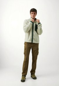 Balta vilnos striukė su tamsiai žaliai užsegama priekyje ir kišene, derinama su rudos spalvos krovinių kelnėmis ir pilkais žygių batais.