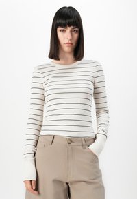 Jonge vrouw met stijl haar, gekleed in een witte blouse met lange mouwen met zwarte strepen en beige hooggehouden broek, staande tegen een witte achtergrond.