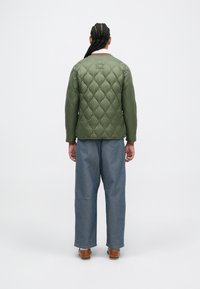 Jachetă verde matlasată cu guler rotund, având modele de diamante ridicate. Asortată cu pantaloni din denim albastru cu picioare largi și pantofi maro.