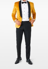 Sametový zlatý blazer s černými saténovými klopami, spárovaný s bílou košilí, černým motýlkem, černými kalhotami a lesklými černými botami.