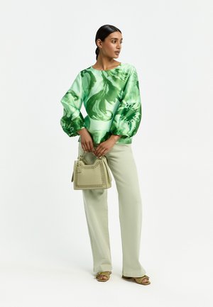 Frau trägt eine grün gemusterte Bluse mit langen Ärmeln, beige weite Hose, grüne Sandalen und hält eine beige Handtasche, steht vor weißem Hintergrund.