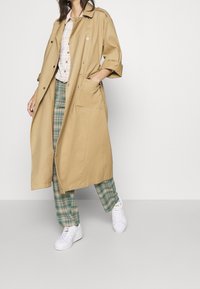 Béžový trenčkot s volnějším střihem, zdobený stříbrnými knoflíky, límcem a bočními kapsami, doplněný o zelené kárované kalhoty a bílé tenisky.