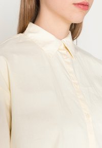 Camisa de botão em tom creme, feita de tecido leve, com colarinho clássico e textura suave. Sem padrões ou detalhes visíveis.