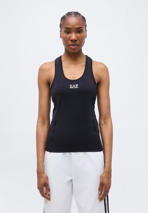 Γυναίκα με πλεξούδες που φοράει μαύρο τοπ EA7 και λευκό παντελόνι με ρίγες στο πλάι, στέκεται μπροστά σε απλό ανοιχτό φόντο.