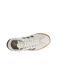 Zapatilla deportiva blanca con rayas negras, parte superior texturizada, suela de goma y cordones planos. Presenta un logo dorado en la lengüeta.