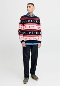 El suéter de punto presenta un diseño en azul marino, rojo y blanco con motivos navideños, patrones a rayas y un escote redondo. Se lleva sobre una camisa de mezclilla.