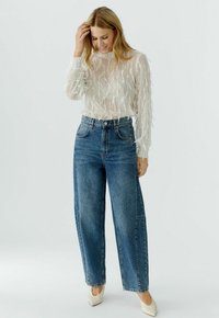 White sheer blouse with textured, wavy detailing; high-waisted blue jeans; white pointed heels; model standing with hand in hair.