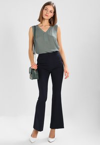 Haut sans manches vert avec un décolleté en V, pantalons évasés bleu marine, talons blancs et un petit sac vert avec des accessoires en or. Tissu lisse et coupe ajustée.