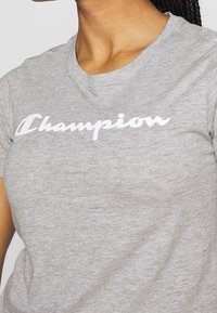 Grå bomulls-t-shirt med korta ärmar, med en vit "Champion"-logotyp över bröstet i kursiv stil. Slät textur.