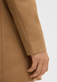 Cappotto in lana beige con una texture liscia, vestibilità su misura e un design pulito e minimale. Il cappotto presenta un leggero risvolto sulla manica.