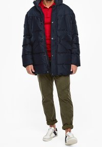 Mørkeblå puffjakke med lynlås og snaplukning, der har to forkert lommer. Båret over en rød polo, parret med olivengrønne bukser og sneakers.