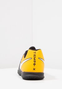 Zapato de fútbol amarillo con acentos negros, parte superior sintética texturizada, suela de goma y marca blanca en el lateral y el talón.