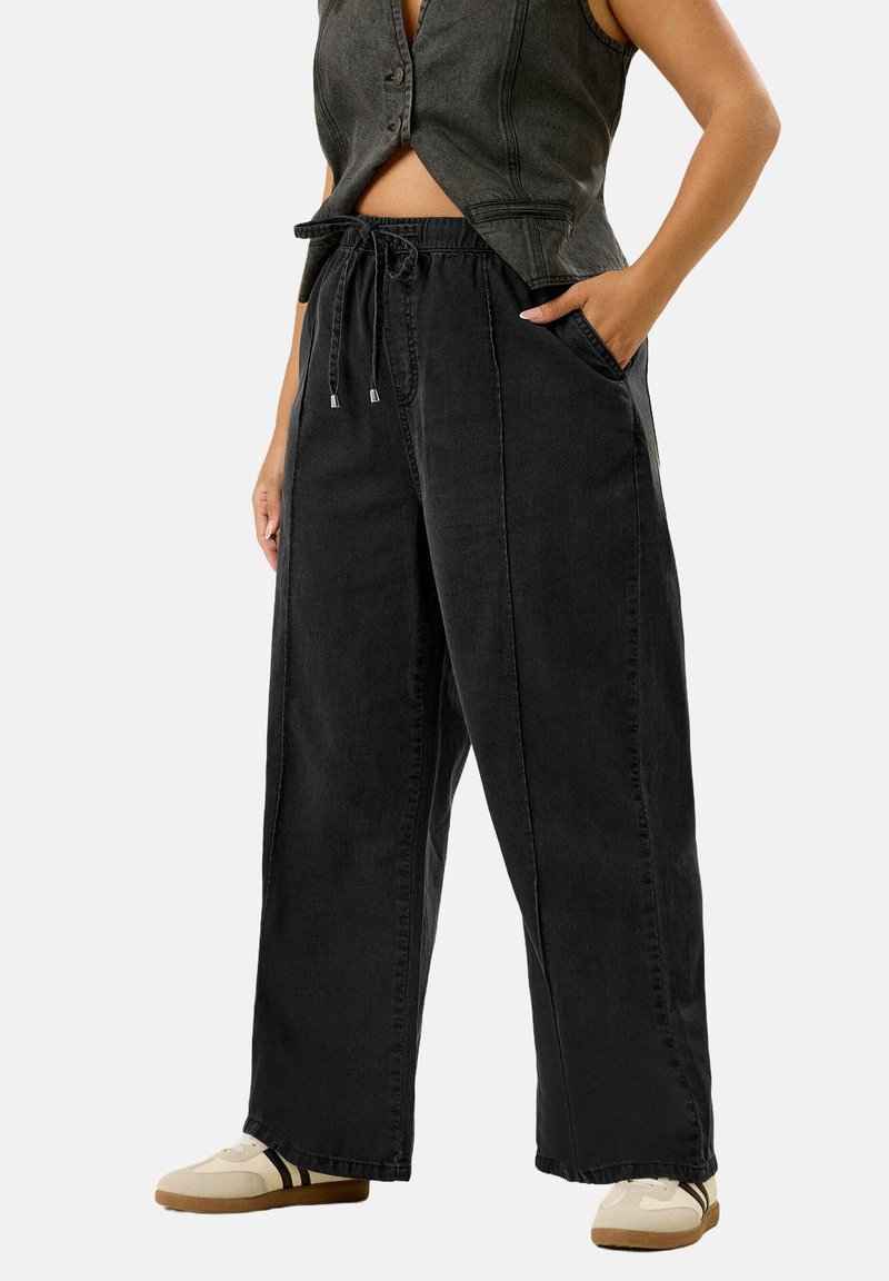 Personne portant un pantalon noir large avec taille à cordon, un gilet sans manches gris fonc�é et des baskets blanches avec des rayures noires.