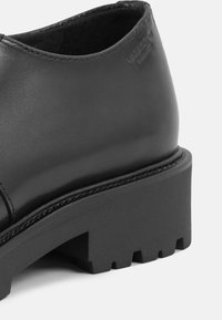 Sapato de couro preto com bico arredondado, sola de borracha espessa e detalhes sutis de costura. Apresenta uma textura suave e minimalismo na marca.