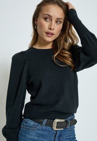 Black knit sweater with ribbed cuffs and shoulder detail, paired with blue denim and a textured black belt featuring a silver buckle.