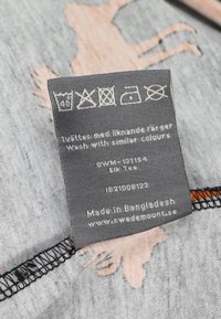 Harmaa kangaslabeli, jossa on hoito-ohjeet ja tyylitelty hirvi-kuvio vaaleanruskeana. Reunoissa on musta ommel. Valmistettu Bangladeshissa.