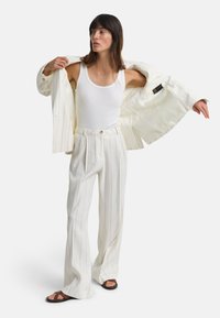 Weiß gestreifter Blazer über einem weißen Tanktop, kombiniert mit weißen, plissierten Hosen. Das Modell trägt braune Riemchensandalen. Glatte Textur.