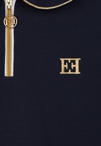 Maglietta zip-up blu navy con colletto bianco, dotata di logo ricamato oro e dettagli in oro sulla zip anteriore. Tessuto morbido.