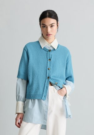 Młoda kobieta ubrana w niebieski, warstwowy, dziergany kardigan, na niebieskiej, paskowanej koszuli i białych spodniach, stojąca na tle jednolitego, szarego tła.