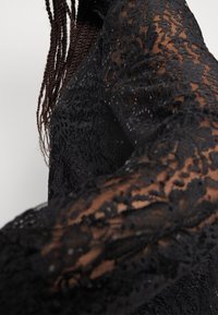 Vêtement à manches longues en dentelle noire avec des motifs floraux et des textures transparentes, présentant des coutures détaillées et des embellissements subtils.