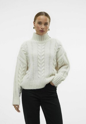 White cable knit sweater with a high neck, featuring textured patterns and ribbed cuffs, paired with black pants.