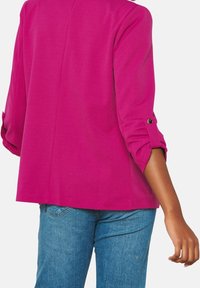 Blazer fucsia con un acabado suave y texturizado, con mangas hasta el codo y un detalle de botón en el puño. Combinado con jeans de mezclilla.