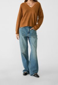 Pull en maille marron à col en V avec manches larges, associé à un jean évasé taille haute bleu clair et des chaussures plates noires en cuir.