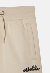 Beige sporta bikses ar ribinātu jostasvietu, auklu, sānu kabatām un melnu logotipa detaļu kreisajā kājā. Mīksta auduma tekstūra.
