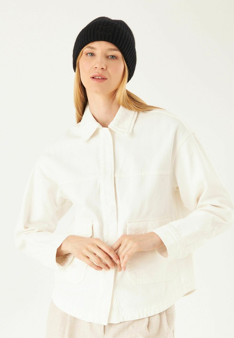 Veste en denim blanche avec un col, des manches longues, deux poches à l'avant et une texture subtile. Le modèle porte un bonnet côtelé noir.