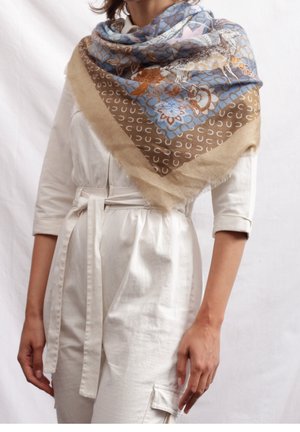Femme portant un foulard à motifs floraux et cerfs en bleu, marron et beige sur une robe crème ceinturée avec des manches retroussées.