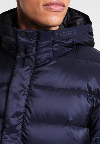 Granatowa kurtka puchowa o połyskującym wykończeniu, z kapturem i zapięciem na zatrzaski. Kołnierz z haftowanym napisem "HOLOGO" w kontrastującym kolorze.