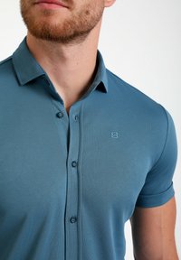 Polo majica s kratkimi rokavi iz mehkega, teksturiranega tkanja v teal barvi. Ima gumbasto zaponko, vezeno ikono in klasično obliko ovratnika.